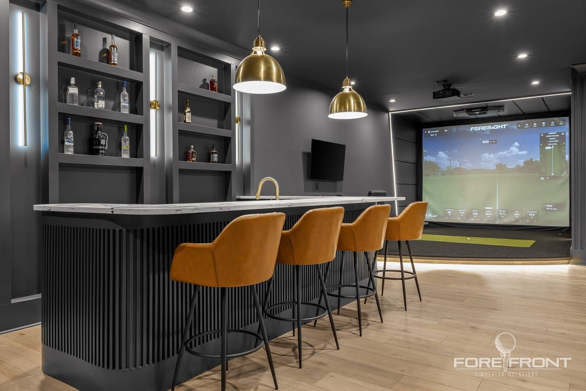 A basement golf simulator bay with a fluted bar, brass pendant lights and leather stools facing the impact screen.